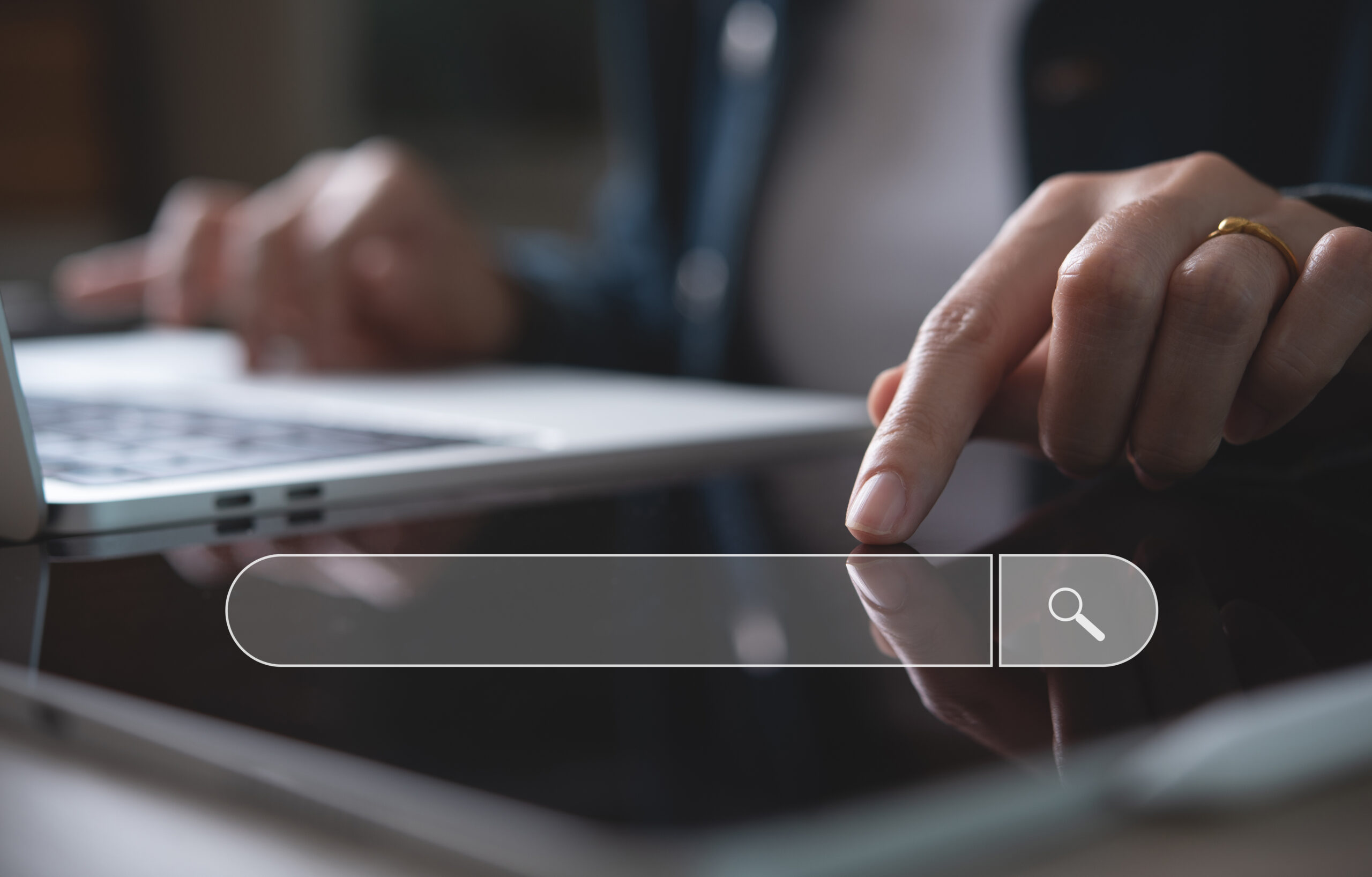 A close-up photograph of a person using a touchscreen device on a dark table. Their index finger touches a semi-transparent search bar overlay equipped with a magnifying glass icon. In the soft-focus background, the person's other hand rests near the keyboard of an open laptop computer.