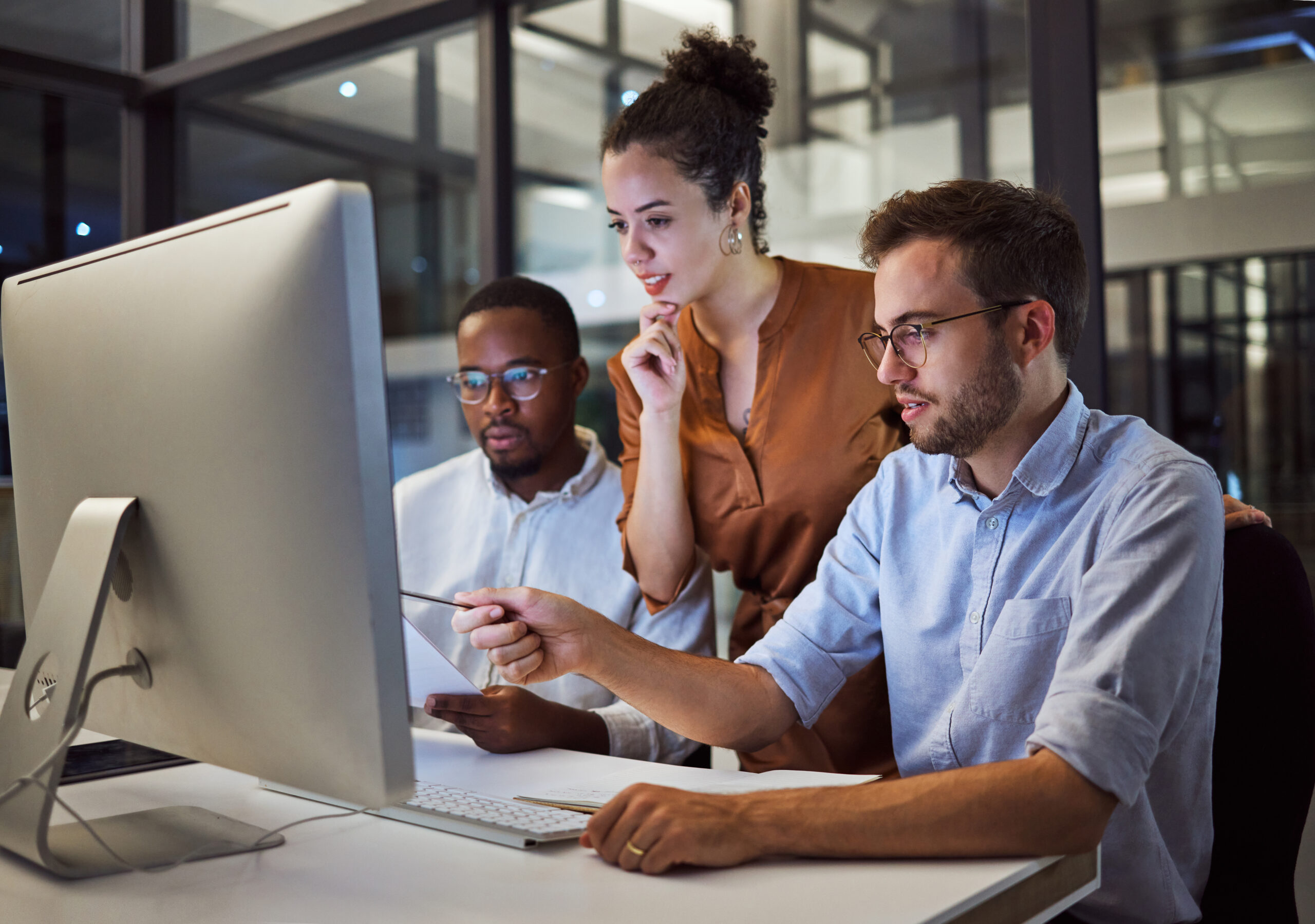 Three diverse colleagues gathered around a desktop computer monitor in a modern office, pointing at the screen and collaborating on a project.