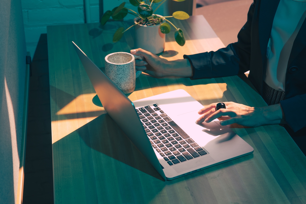 High-angle shot of a person at a wooden desk using a silver laptop. One hand holds a ceramic mug while the other, wearing a prominent black ring, operates the trackpad. Sunlight streams across the desk creating geometric shadows, set against moody teal lighting and a potted plant in the background.